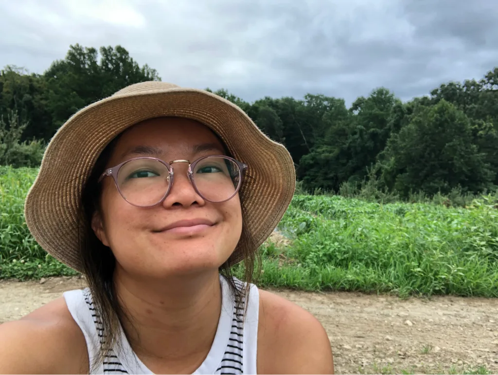 K.J. Chien smiles at the camera while outdoors, wearing a wide-brimmed straw hat, round glasses, and a sleeveless striped shirt. Behind them is a dirt path and lush green vegetation with dense trees under a cloudy sky.