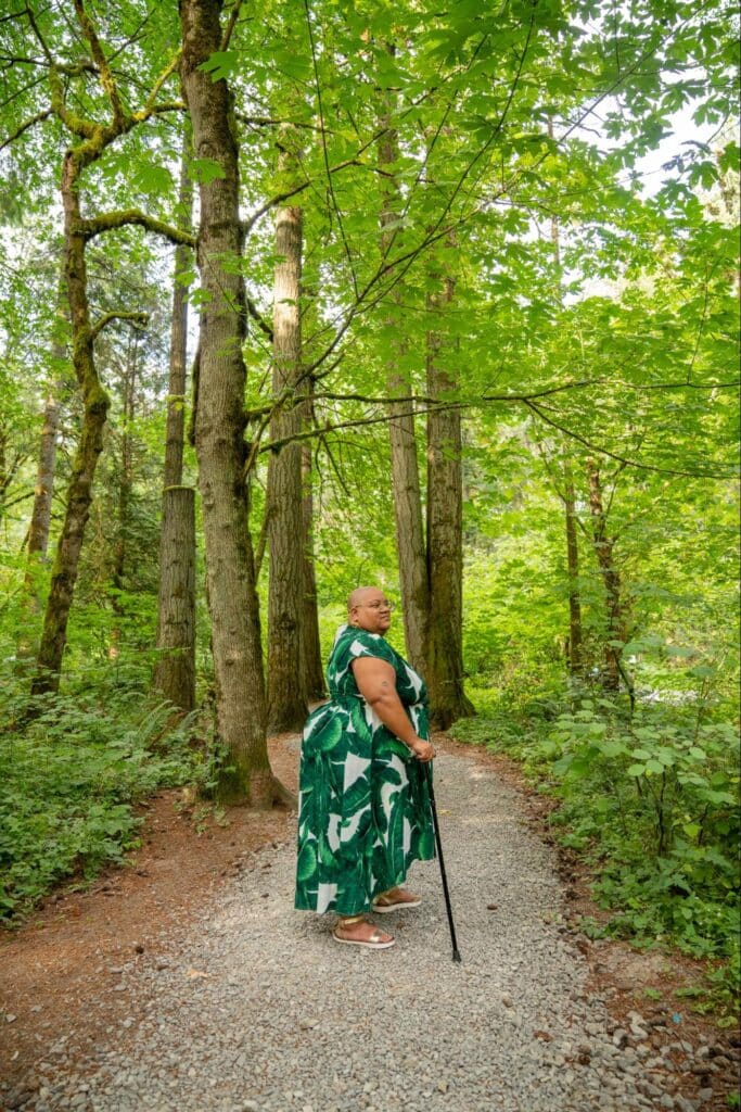 Side portrait shot of a Black non-binary person standing regally in a rocky forest grove with their cane, their shaved head framed by lush greenery that gives the illusion of a headpiece. They wear glasses, a long flowing leaf print dress, and gold sandals. Side portrait shot of a Black non-binary person standing regally in a rocky forest grove with their cane, their shaved head framed by lush greenery that gives the illusion of a headpiece. They wear glasses, a long flowing leaf print dress, and gold sandals.