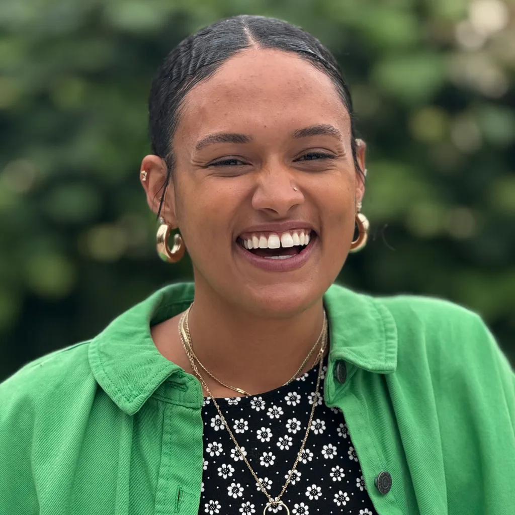 A woman with dark hair pulled back smiles brightly. She wears gold hoop earrings, layered necklaces, a green shirt over a black-and-white floral top, and stands in front of a blurred green outdoor background.